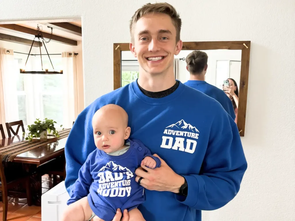 Father holding his baby son, both smiling in matching “Adventure Dad” and “Dad’s Adventure Buddy” sweatshirts, symbolizing intentional and joyful parenting